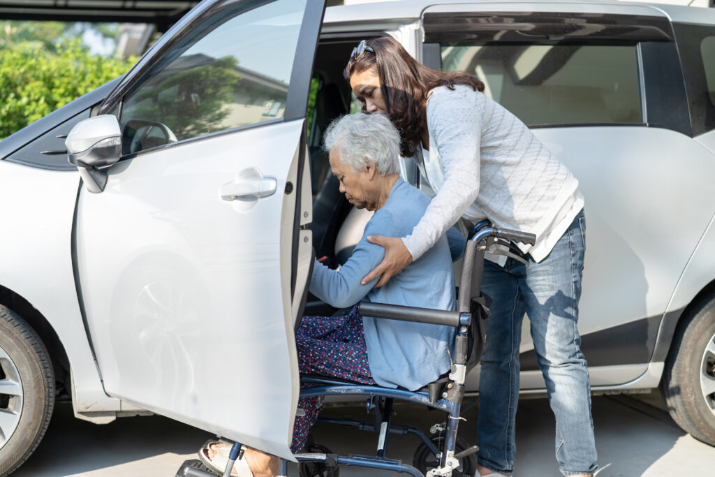 A caregiver assists an elderly woman from a wheelchair into a car.