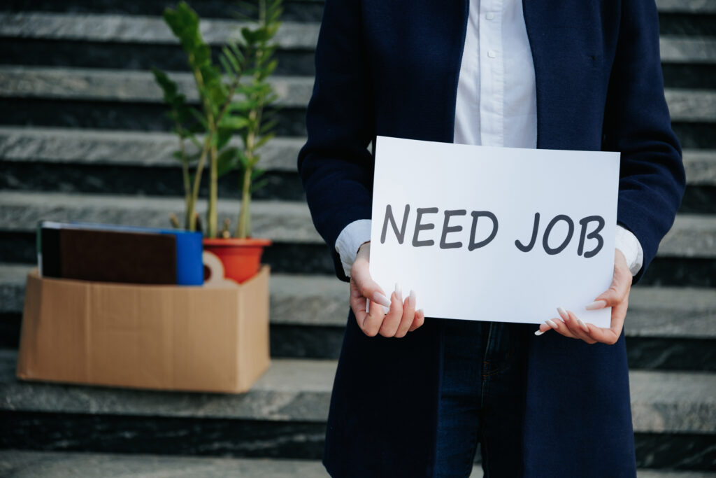 Person holding a sign that says 'NEED JOB' near packed boxes.